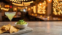 Cocktail with guacamole dip and tortilla chips on wooden board against warm golden bokeh lights, perfect for bar, restaurant, and hospitality marketing.