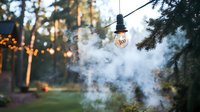 Glowing light bulb hanging outdoors with atmospheric smoke and bokeh lights in evening forest setting, creating magical ambiance for garden party decoration.