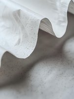 Elegant white fabric with gentle wave-like folds creates soft shadows and texture, scattered with tiny dark speckles against a neutral gray background. Minimalist composition.