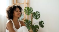 Young African American woman in white top practicing mindfulness meditation at home with monstera plants in bright modern interior space.