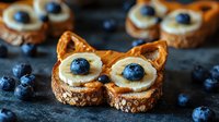 Creative breakfast toast shaped as cat face with banana slices, blueberries and peanut butter on multigrain bread, scattered fresh berries on dark background.