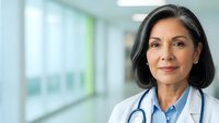 Professional Hispanic female doctor with stethoscope smiling confidently in modern hospital corridor wearing white medical coat for healthcare marketing.