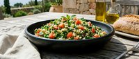 Tabbouleh salad with fresh parsley, tomatoes, and bulgur wheat in black bowl on outdoor table with bread and olive oil.
