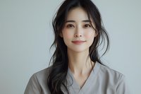 Young Asian woman with long dark hair wearing gray professional attire smiling warmly against neutral background, natural makeup and gentle expression for wellness and beauty concepts.