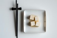 Fresh tofu cubes served on white ceramic plate with black chopsticks on clean background for healthy Asian cuisine and vegetarian cooking concepts.