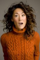 Asian woman with surprised expression wearing orange turtleneck sweater, curly brown hair flowing. Studio portrait capturing genuine emotion and style for advertising and lifestyle content.