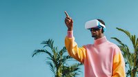 Young man wearing VR headset with pink and yellow sweater gesturing upward against blue sky, surrounded by tropical palm plants.
