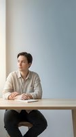 Young Caucasian businessman in beige shirt sitting at wooden desk looking thoughtfully away in modern minimalist office space with neutral background.