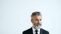 Mature male professional with gray hair and beard wearing dark business suit and tie against white background, suitable for corporate portraits and business communications.