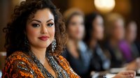 Confident Hispanic businesswoman with curly hair and elegant jewelry at corporate meeting, colleagues blurred in background.