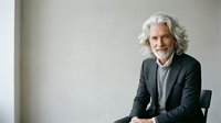Mature man with white hair and light skin tone, wearing dark blazer over gray shirt, seated against neutral background, professional portrait for business and corporate use.