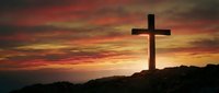 Christian cross silhouette against dramatic sunset sky with orange and red clouds on rocky hilltop for religious and spiritual themes.
