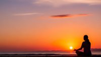 Woman silhouette meditating in lotus pose on beach during golden sunset with orange and purple sky colors reflecting peaceful mindfulness practice by ocean.