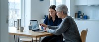 Two professional women collaborating on laptop computer in modern office workspace discussing business strategy and data analysis.