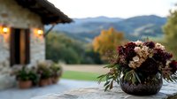 Moody floral arrangement with dark burgundy and cream flowers in rustic vase against blurred Tuscan villa background, warm evening lights and rolling hills landscape.