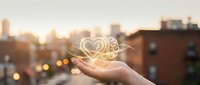 Hand holding glowing heart with decorative swirls against blurred city skyline at sunset, symbolizing love, care, compassion and human connection in urban environment.