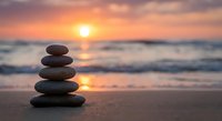 Balanced stone stack on beach at sunset with golden sky and ocean waves creating peaceful zen meditation atmosphere for wellness and mindfulness practices.