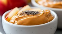 Creamy peanut butter topped with chia seeds in white ceramic bowl, selective focus on smooth texture and garnish, fresh apple slices in background.