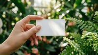 Female hand holding blank white business card against lush green tropical garden background, mockup for branding and design.