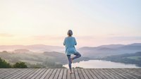 Senior woman in blue shirt practicing yoga on wooden deck overlooking misty valley landscape with lake and mountains at sunrise, wellness and mindfulness concept.