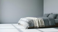 Minimalist bedroom interior with unmade bed featuring gray and white bedding, cream throw blanket, and pillows against white wall with natural light for home decor and interior design projects.