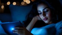 Young Hispanic woman lying on couch at night, illuminated by blue light from digital tablet screen, with bokeh lights in background creating moody atmosphere.