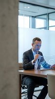 Middle-aged Caucasian male professional wearing business suit and glasses working on tablet in modern glass-walled office conference room with natural light, suitable for corporate, technology, and business concepts.