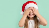 Happy young Caucasian girl wearing red Santa hat covering eyes with hands while smiling during Christmas celebration on mint green background.