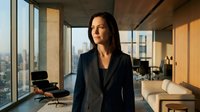 Middle-aged woman with long dark hair and East Asian ethnicity wearing dark business blazer standing in modern high-rise office with floor-to-ceiling windows overlooking cityscape. Professional corporate environment.