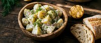 Creamy potato salad with fresh herbs and roasted garlic served in wooden bowl with crusty bread slices on rustic dark table for food styling and culinary content.