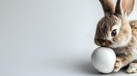 Adorable brown bunny rabbit with long ears holding white egg against neutral gray background, perfect for Easter holiday designs and spring celebrations.