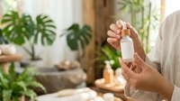 Young woman with medium skin tone holding a white cosmetic bottle in a bright, plant-filled interior space. Suitable for beauty, skincare, wellness and natural product marketing campaigns.