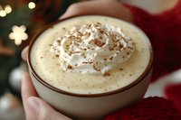 Creamy hot chocolate topped with whipped cream and cocoa powder in ceramic mug, held against festive holiday background with bokeh lights and decorations.
