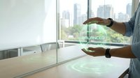 Male professional with medium skin tone wearing business casual attire demonstrating augmented reality interface with holographic digital elements and data visualization on table in modern office with city skyline view.
