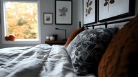 Cozy bedroom interior with unmade bed, gray and brown textured bedding, botanical wall art, and window with autumn view creating peaceful morning atmosphere.