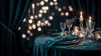 Elegant holiday table setting with teal velvet tablecloth, crystal glasses, lit candles and warm bokeh lights creating festive dining atmosphere.
