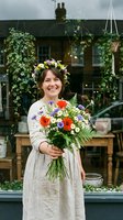 Young woman with dark hair and fair complexion wearing a floral crown and beige linen dress, holding a vibrant bouquet of red poppies, white daisies, and purple flowers with green foliage in front of a rustic garden shop with hanging ivy planters.