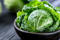 Fresh savoy cabbage head with crisp green leaves in dark ceramic bowl on wooden table, close up view with natural light and selective focus.