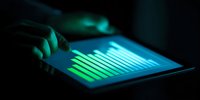 Person analyzing financial data on tablet with glowing green charts in dark environment, monitoring market trends for investment decisions.