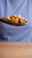 Mixed nuts and dried fruits on wooden spoon against purple fabric background. Healthy snacking, cooking ingredients, natural nutrition concept.