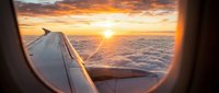Airplane window view of aircraft wing during golden hour sunset flight above clouds and ocean landscape.
