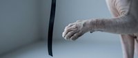 Senior man with wrinkled skin and age spots extends arm toward black curved object in minimalist studio setting, conveying reaching, aging, or interaction themes.
