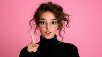 Young Asian woman with messy bun hairstyle wearing glasses and black turtleneck pointing up with finger against pink background, expressing bright idea or solution.