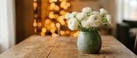 White roses in green ceramic vase on wooden table with warm bokeh lights creating cozy home decor atmosphere for interior design and lifestyle.