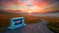 Clean energy for all neon sign glowing on remote mountain road at sunset, winding asphalt path through golden moorland landscape with dramatic orange sky.