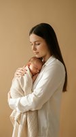 Young mother with long dark hair tenderly holding sleeping newborn baby wrapped in beige blanket against warm neutral background for maternity concepts.