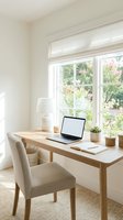 Bright home office workspace with wooden desk, laptop, and upholstered chair positioned near large windows overlooking garden, featuring white lamp and potted plants for work or study.