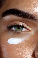 Close-up of green eye with shimmering golden eyeshadow and white moisturizer streak on glowing, dewy skin. Macro beauty shot highlighting skincare and cosmetics.
