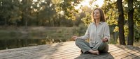 Middle-aged Caucasian woman practicing mindfulness meditation outdoors on wooden deck during golden hour sunset with peaceful forest background.