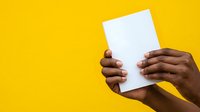Person with dark skin tone holding blank white card against bright yellow background for messaging or advertising purposes.
